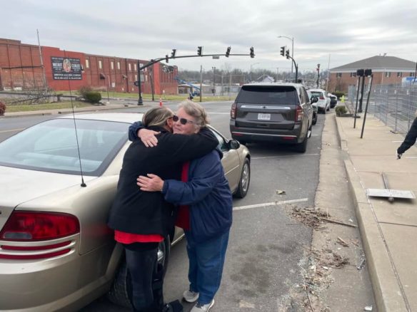 Two women hugging in front of a car parked on the side of a street.