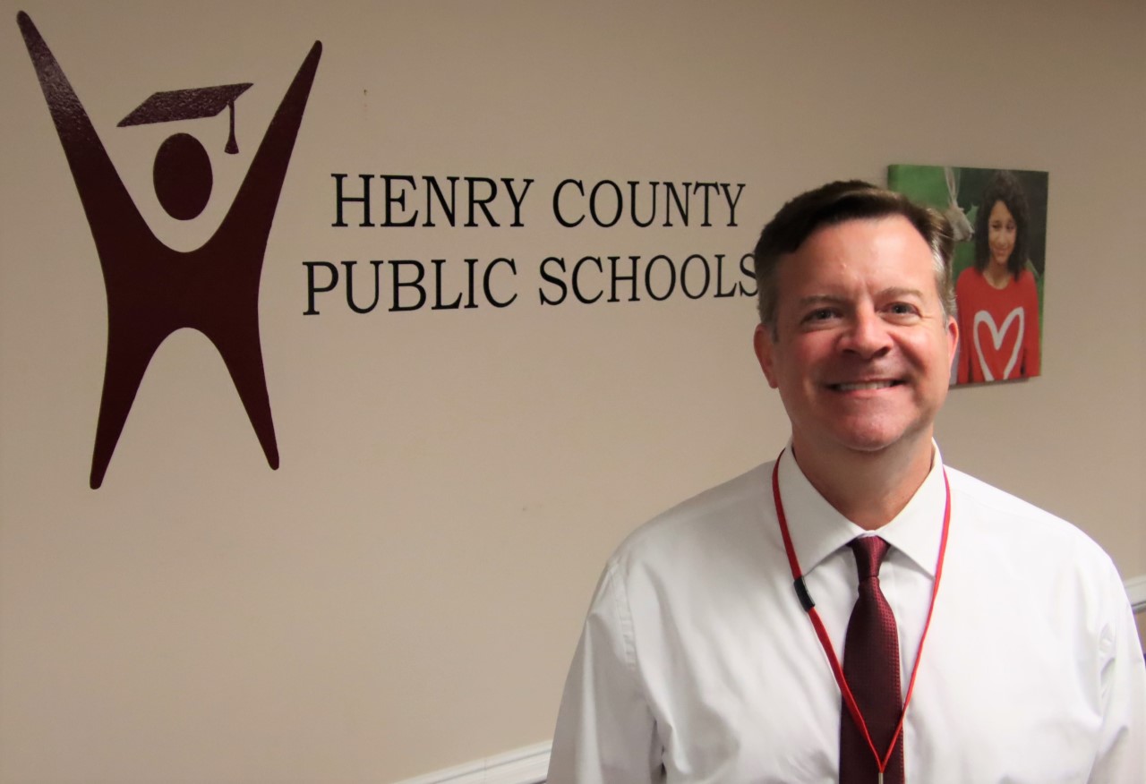 A man smiling and standing in front of a sign on a wall that reads: Henry County Public Schools
