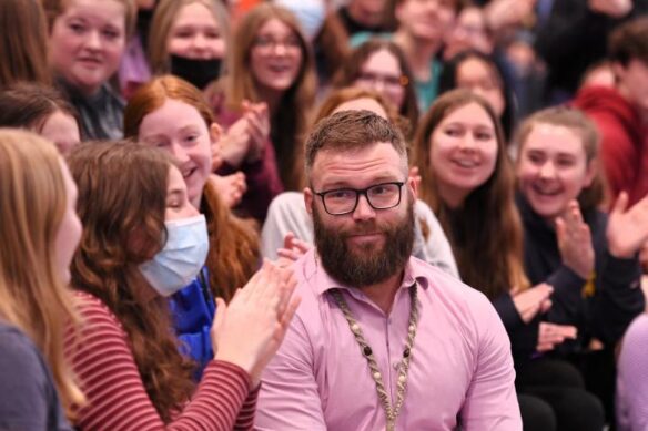 A man smiling softly while students around him smile and clap.