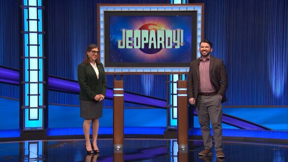 A woman and a man standing in front of a large screen that reads: Jeopardy! 