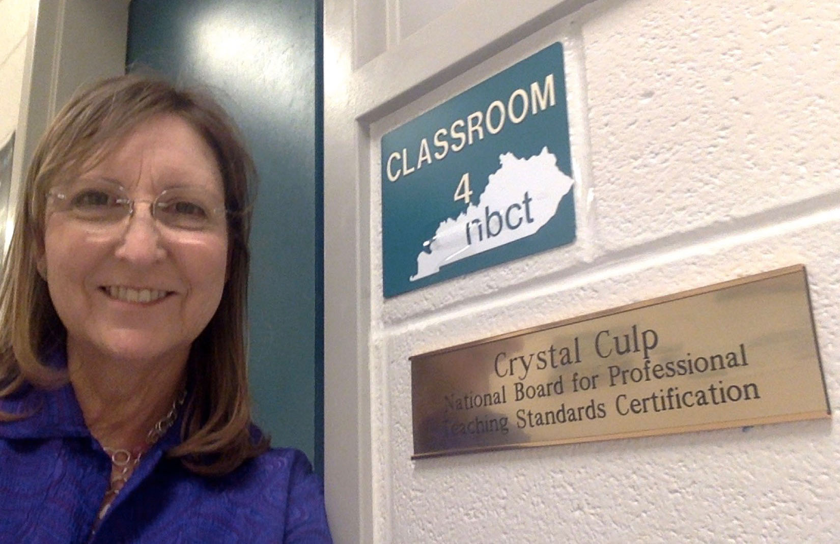 A woman smiling next to two plaques on a wall that read: " Classroom 4 NBCT" and "Crystal Culp National Board for Professional Teaching Standards Certification"