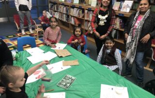 A group of parents and students gathered around a table with crafts on it. There are books on shelves behind them.