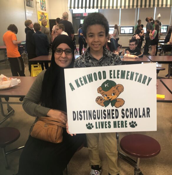 A woman and child smile as they hold a sign that reads: "A Kenwood Elementary Distinguished Scholar Lives Here" with a cartoon bear on it. 