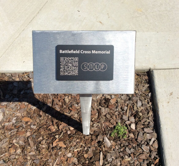 A photo of a silver plaque in the ground that has a QR code on it and reads: Battlefield Cross Memorial.