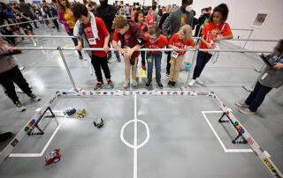 Students watch a robotic soccer game on the floor.