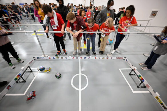 Students watch a robotic soccer game on the floor. 