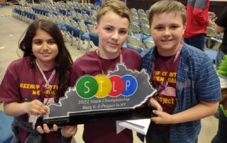 Three students hold a trophy in the shape of the state of Kentucky that reads: STLP, 2022 State Championship, Best K-5 Project in Kentucky.