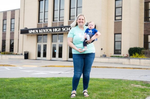 A woman holds a child in front of a building with a sign that reads: Simon Kenton High School. 