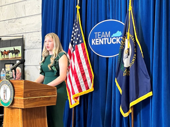 A student stands in front of a U.S. flag and Kentucky state flag and a sign that reads "Team Kentucky" as she speaks from behind a podium.