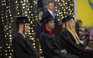 Picture of three students sitting on a stage wearing wearing graduation caps and gowns while somebody speaks in the background.