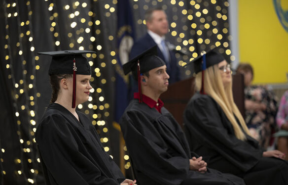 Picture of three students sitting on a stage wearing wearing graduation caps and gowns while somebody speaks in the background.