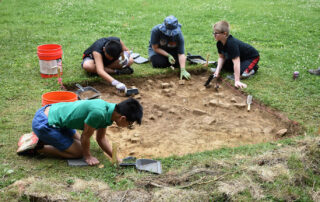A picture of four students sitting on the ground, excavating a small square of ground using shovels and brushes.