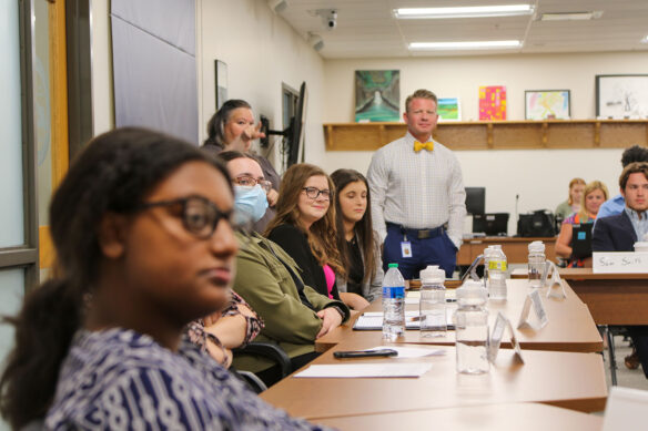 Picture of several students sitting at long table, listening to a speak who is off screen.