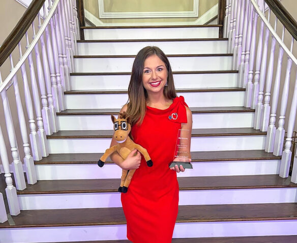 A woman stands smiling in front of a staircase holding a trophy and a stuffed horse. 