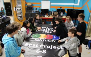Students sit around a banner on a table that reads: State Champions 2022 STLP Kentucky, Best Project, 6-8.