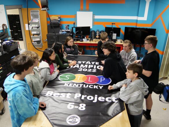 Students sit around a banner on a table that reads: State Champions 2022 STLP Kentucky, Best Project, 6-8.