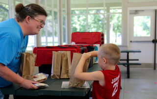 Picture of a young boy reaching into a brown lunch bag while a woman across the table smiles at him.