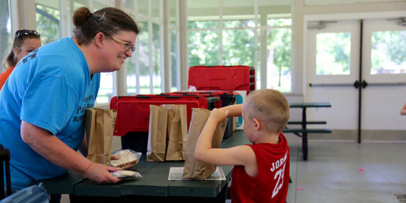 Picture of a young boy reaching into a brown lunch bag while a woman across the table smiles at him.