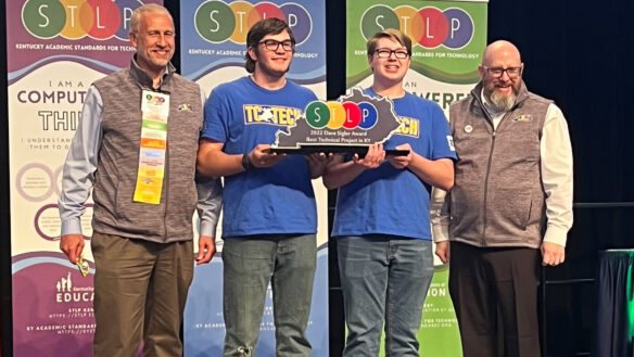A photo of four people standing smiling. The middle two people are holding a trophy in the state of Kentucky that reads: "STLP."