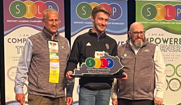 A photo of three people standing and smiling. The person in the middle is holding a trophy in the shape of the state of Kentucky that reads: "STLP."