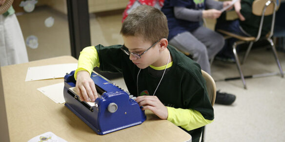 Picture of a young boy reading what he has typed on a Braille typewriter.