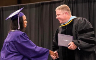A student wearing a graduation cap and gown shakes hands with a man handing her a diploma.