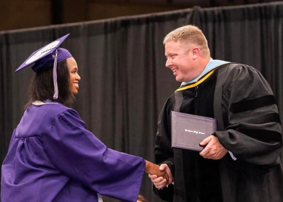 A student wearing a graduation cap and gown shakes hands with a man handing her a diploma. 