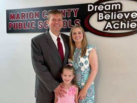 A man and woman stand smiling with their child in front of a sign that reads: Marion County Public Schools, Dream, Believe, Achieve