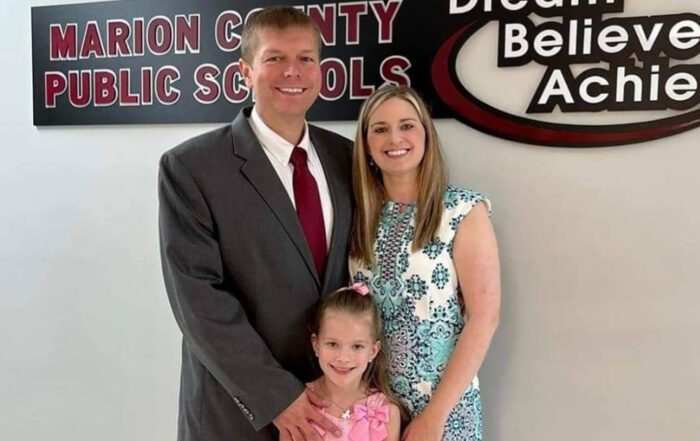 A man and woman stand smiling with their child in front of a sign that reads: Marion County Public Schools, Dream, Believe, Achieve
