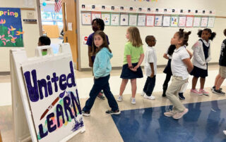 Students walking in a hallway past a sign that reads: "United We Learn"