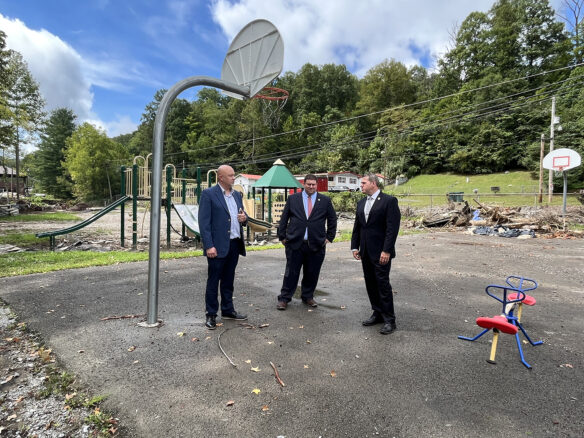 Three men stand on a damaged outdoor basketball court in front of debris and a damaged playground. 