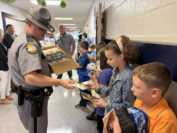 A picture of a state trooper handing out books to students in a school hallway.