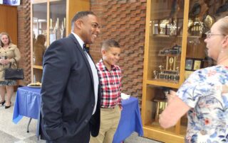 Danville Independent Superintendent Greg Ross stands next to a young boy while talking to a woman. They stand in front of a trophy case in a school.