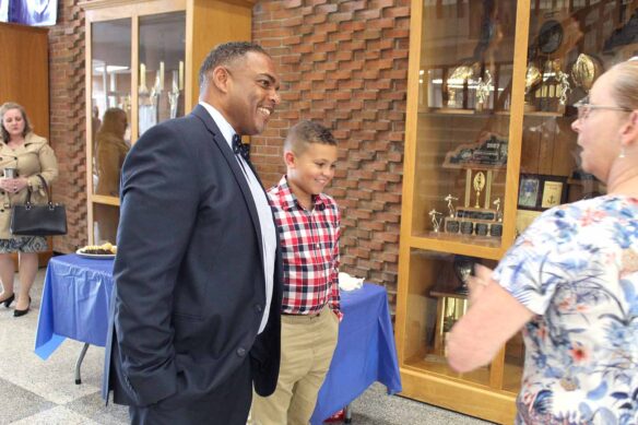 Danville Independent Superintendent Greg Ross stands next to a young boy while talking to a woman. They stand in front of a trophy case in a school. 