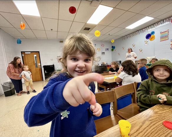 A smiling student points at a camera. She is in a classroom. 