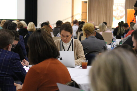 Photo focused on a woman working on a laptop at the 2022 Continuous Improvement Summit. She is surrounded by others sitting at tables. 