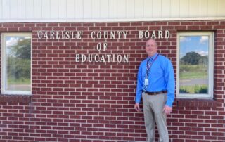 Casey Henderson stands in front of a brick wall that says: Carlisle County Board of Education.