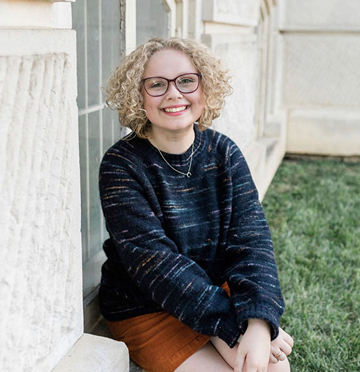 Picture of a young woman sitting outside, wearing glasses and smiling.