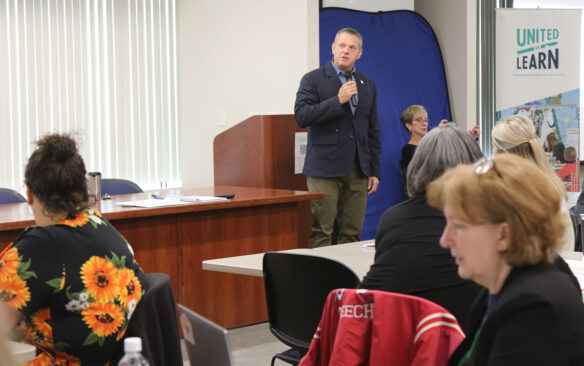 A photo of a man presenting to a room full of people. A banner behind him reads United We Learn.