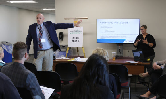 Picture of a man in a suit pointing to a screen and talking to a group of people.