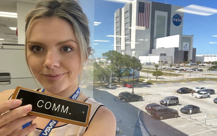 A picture of a woman taking a selfie in front of a window holding a nametag that says comm. In the background is a large building with the NASA logo.