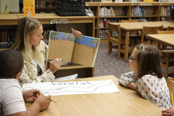A picture of a college student reading a book to two elementary students sitting in a library.