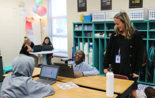 Picture of an elementary school teacher laughs with her student. The student is sitting at a desk, working on a laptop.