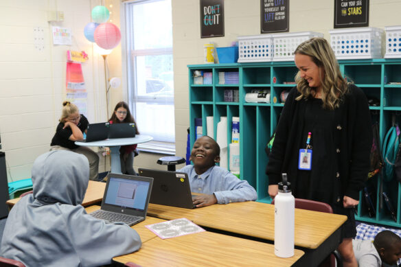 Picture of an elementary school teacher laughs with her student. The student is sitting at a desk, working on a laptop.