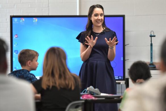 Picture of a woman talking excitedly with her hands to a classroom of students. A large touchscreen monitor is behind her.