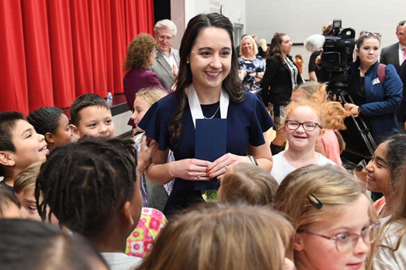 A picture of a woman standing in a school gymnasium holding a small envelope with students crowding around her smiling and laughing.