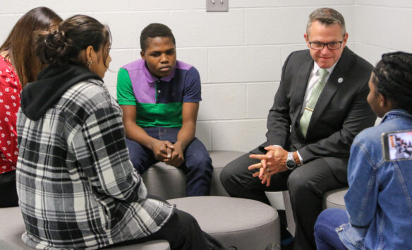 Education Commissioner Jason E. Glass sits with three students at a round chair area. 