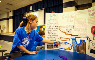 A girl points to a poster board describing a project about elk.
