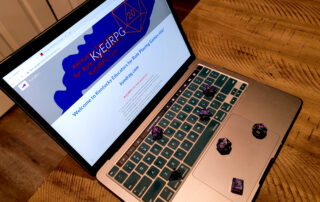 Seven die used for role-playing games are displayed across the keyboard of a laptop, which has the KyEdRPG website loaded on the monitor and is sitting on a wooden table.