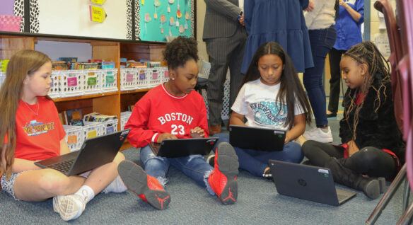 A group of four students sit on the floor of a classroom while they work on laptops.
